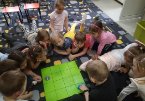 Grupka dzieci siedzi na dywanie dookoła kwadratowej zielonej planszy. Obserwują jak porusza się po kolejnych kwadratach robot-mysz.